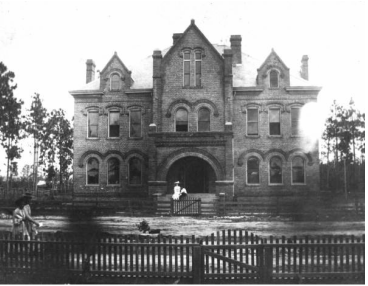 Historic brick building with ornate architecture, featuring multiple gables and arched windows, surrounded by a wooden fence and trees.