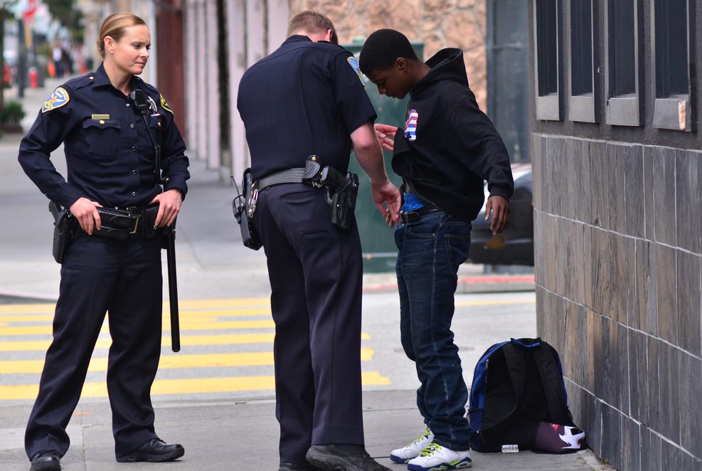 Two police officers are interacting with a young man on the street, one officer is searching him while the other observes. A backpack is placed on the ground nearby.