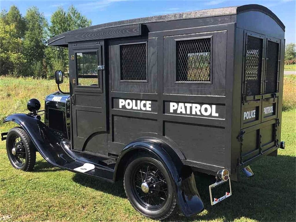 A vintage black police patrol car with large white 'POLICE PATROL' markings on the sides, parked on grass with trees in the background.