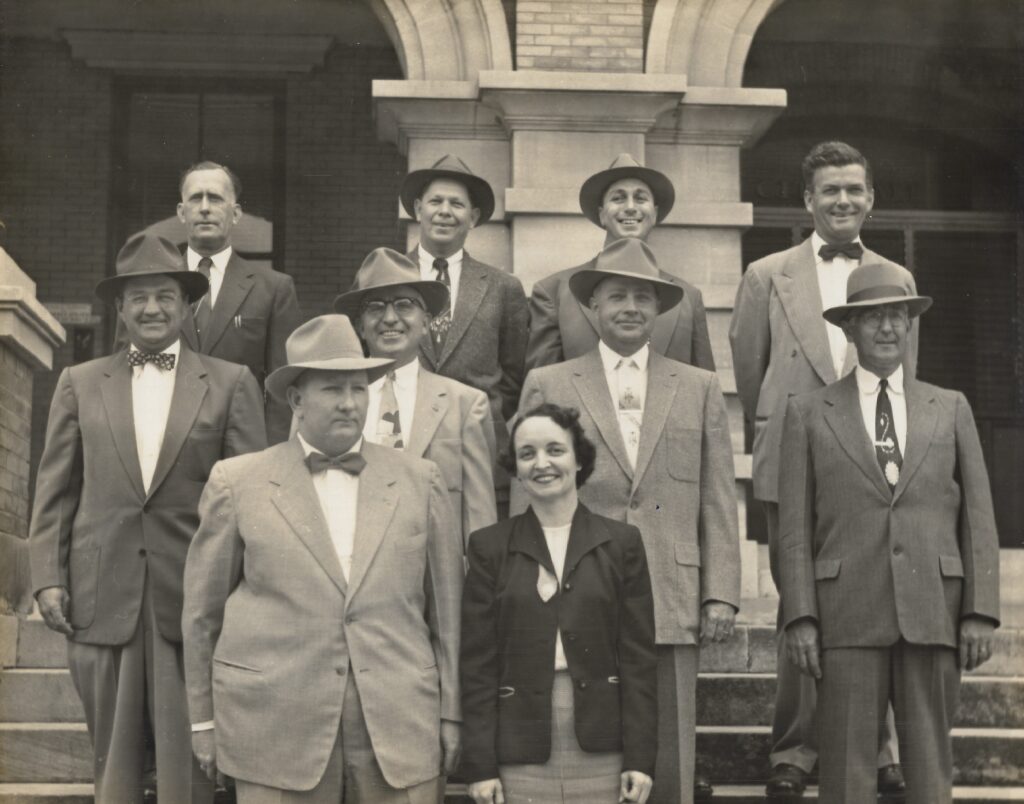 A black and white photo of a group of eleven people, including a woman, standing on the steps of a building. Most individuals are wearing suits and hats, with several showing bow ties. The group poses for the camera with a serious yet cordial demeanor.