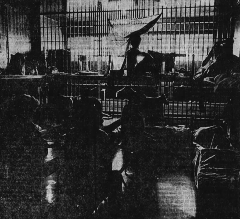 Dimly lit interior of a prison or detention facility, featuring barred cells and figures sitting on the floor with bedding.