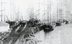 Historic black and white photograph of a busy dock with multiple sailing ships and a wooden pier.