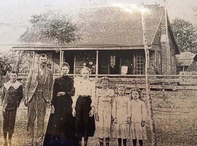A historic black and white photo of a family standing in front of a wooden house, featuring five children and two adults.