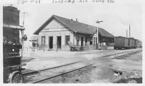 Historic black and white image of a train station with wooden architecture, surrounded by railway tracks and a train car.