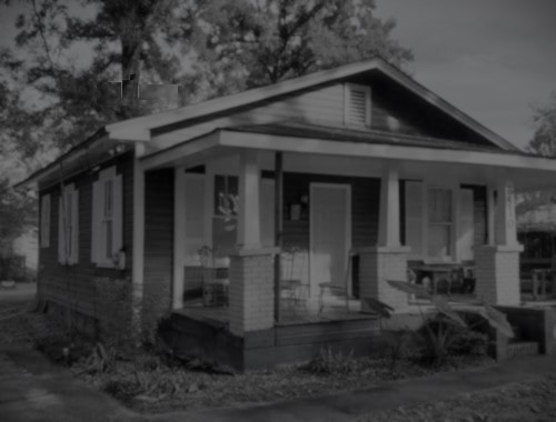 A black and white image of a small, single-story house with a front porch, featuring white columns and windows.