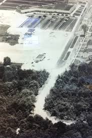 Aerial view of a flooded area with buildings, roads, and a swimming pool visible. The surrounding landscape includes dense trees.