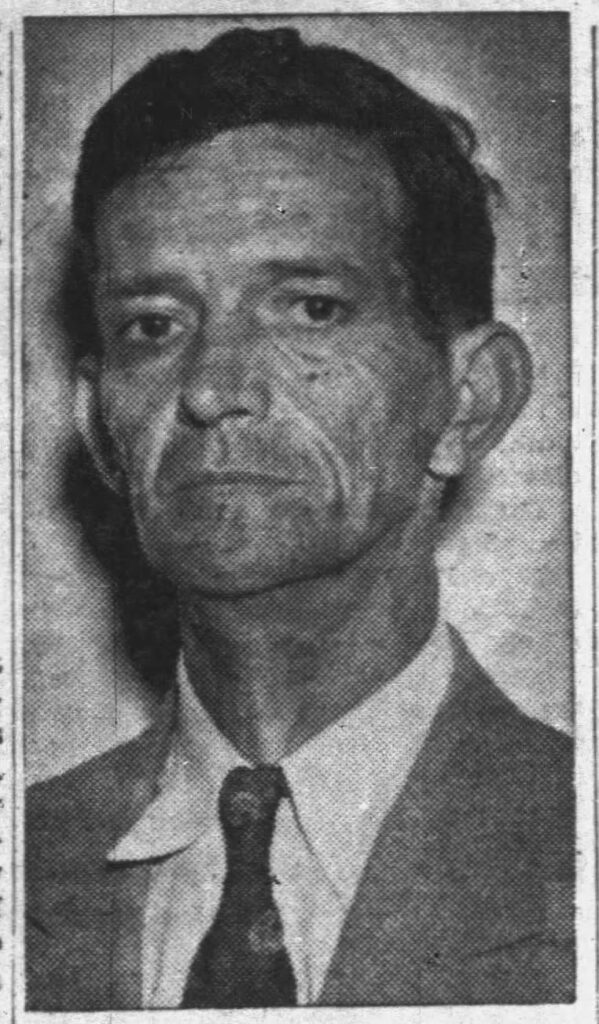 A black and white portrait of a middle-aged man with a serious expression, wearing a suit and tie. The background appears plain.