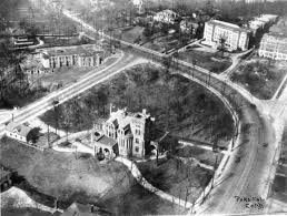 Aerial view of a historical building surrounded by roads and greenery, showcasing a curved road and several nearby structures.