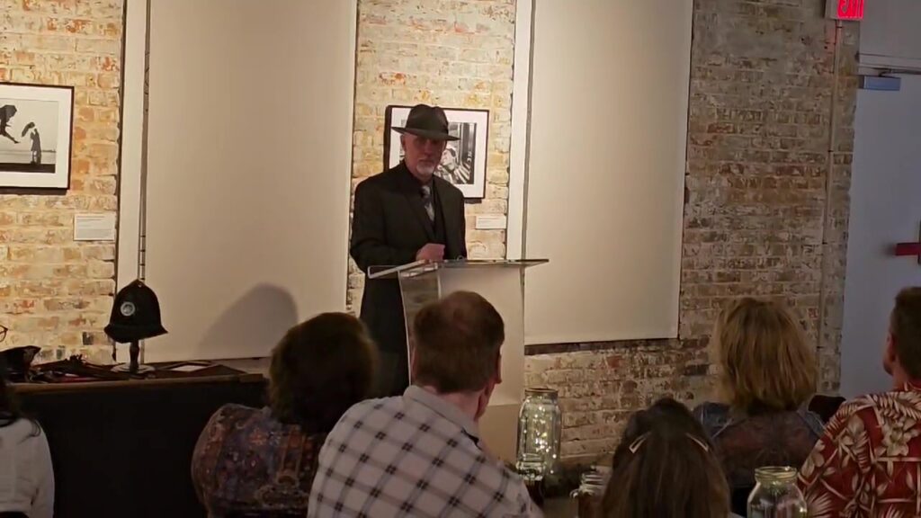 A speaker in a hat presenting at a podium in an art gallery, with an audience visible in the foreground and artwork hanging on the brick walls.