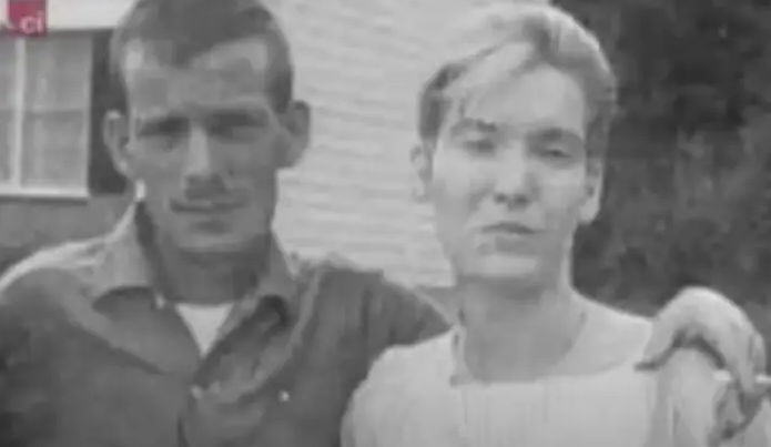 Black and white photograph of a man and woman standing close together, with the man wearing a button-up shirt and the woman in a light dress, both appearing serious.