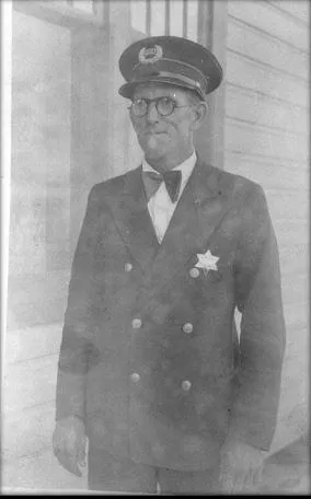 A man in a military-style uniform with a cap, glasses, and a star badge, standing against a wall.