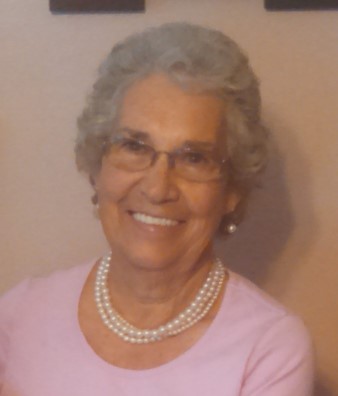 An elderly woman with short, curly gray hair smiles while wearing glasses and a pearl necklace, dressed in a pink top.