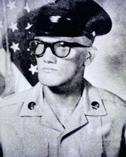 A black and white portrait of a soldier wearing a military uniform and cap, with glasses, standing in front of a US flag.