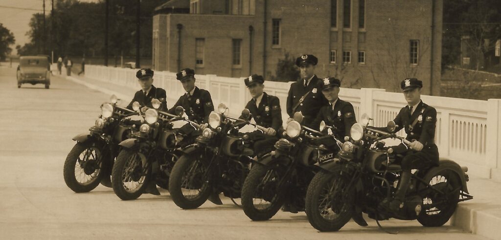 A historic black and white photo of five police officers on motorcycles, lined up on a street. The officers are wearing uniforms and hats, with one officer standing among them. A vintage car is visible in the background, along with a building.