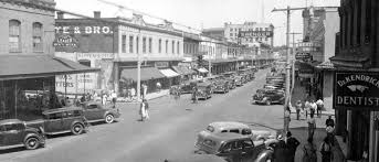 Black and white historic street view showing cars parked along a bustling commercial street with various shops and buildings.