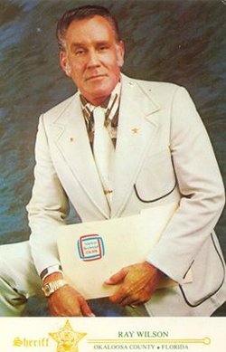A man dressed in a white suit with a badge on his lapel, sitting and holding a document, representing a sheriff's position in Okaloosa County, Florida.