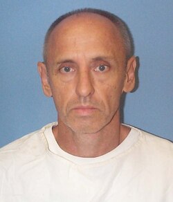 Mugshot of a man with a bald head and serious expression, wearing a white shirt against a blue background.