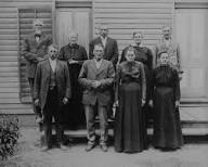 A historical black and white photograph of a group of eleven people posed in front of a wooden building, dressed in early 20th-century clothing.