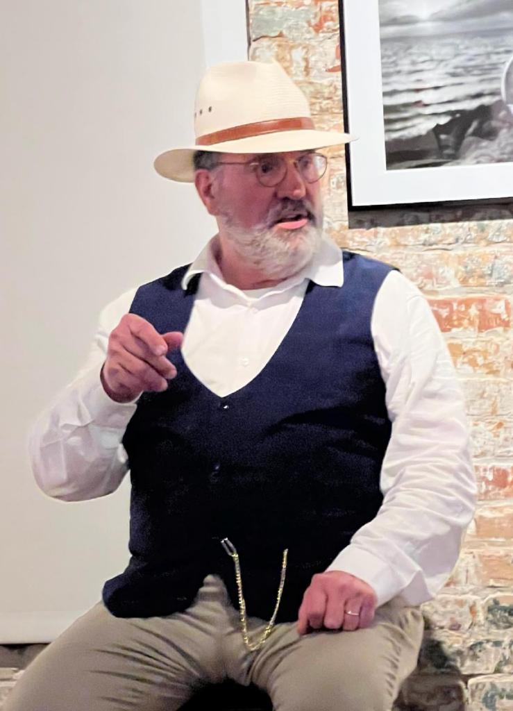 A man with a beard and glasses wearing a straw hat, white shirt, and navy vest, seated and gesturing while speaking in front of a brick wall.