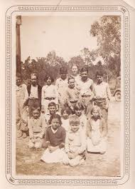 A historical black and white photo of a group of children posing together outdoors, with some standing and others sitting. Trees and vintage vehicles are visible in the background.