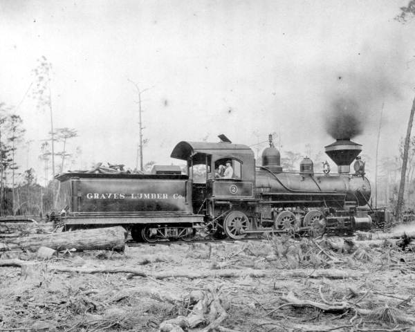 Black and white historical photo of a steam locomotive with 'Graves Lumber Co' on the side, set in a wooded area with felled trees.