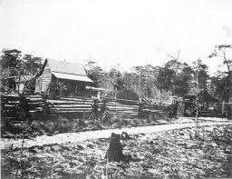 Black and white photograph of a rustic cabin surrounded by a wooden fence and trees, with a dirt path leading to the structure.
