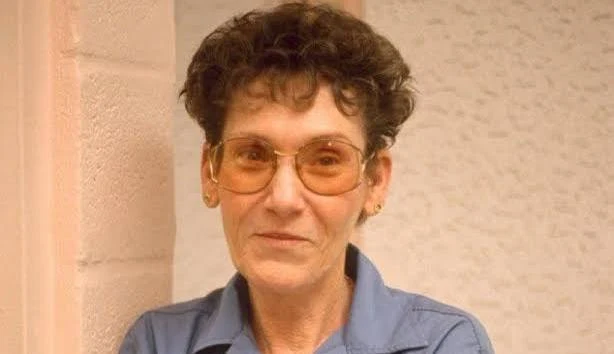 An older woman with short curly hair wearing large sunglasses and a blue shirt, standing against a textured wall.