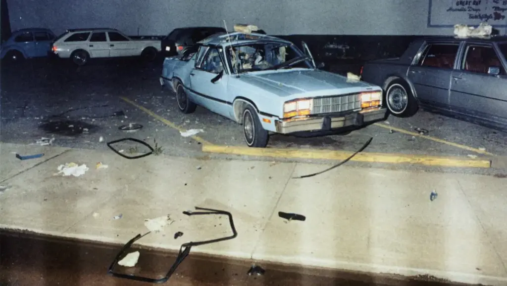A damaged blue sedan parked in a lot, surrounded by debris and other vehicles. The car shows signs of significant wear, with broken windows and trash scattered on the ground.