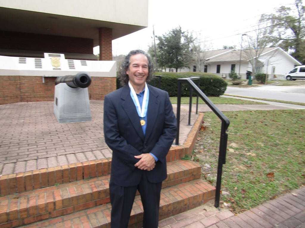 A man with long curly hair wearing a dark suit and a medal stands smiling in front of a historic cannon displayed on a brick platform.
