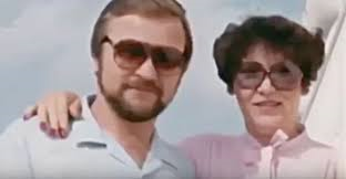 A man and woman posing together outdoors, both wearing sunglasses. The man has a beard and is dressed in a light blue shirt, while the woman is in a pink blouse. They appear to be smiling and enjoying their time.