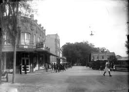 Historic street scene with pedestrians and vintage buildings, featuring shops and a canopy of trees in the background.