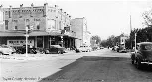 Black and white photograph of a historic downtown street showing parked cars, storefronts, and early 20th-century architecture.