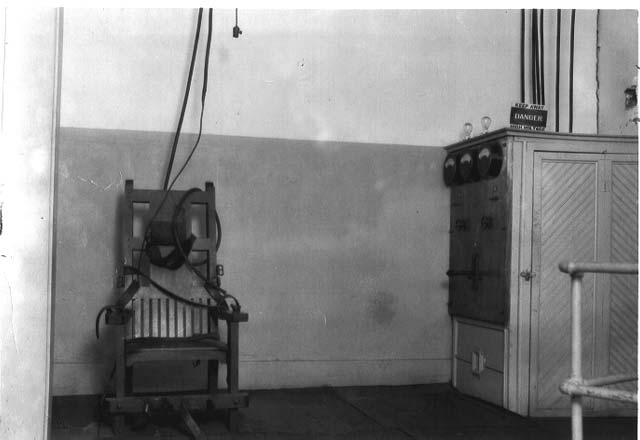 A black and white photograph of an electric chair in a room, with a device next to it. The chair is equipped with straps and a head restraint.
