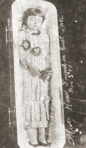 A historic photograph of a young girl lying in a glass casket, dressed in a white gown and holding flowers.