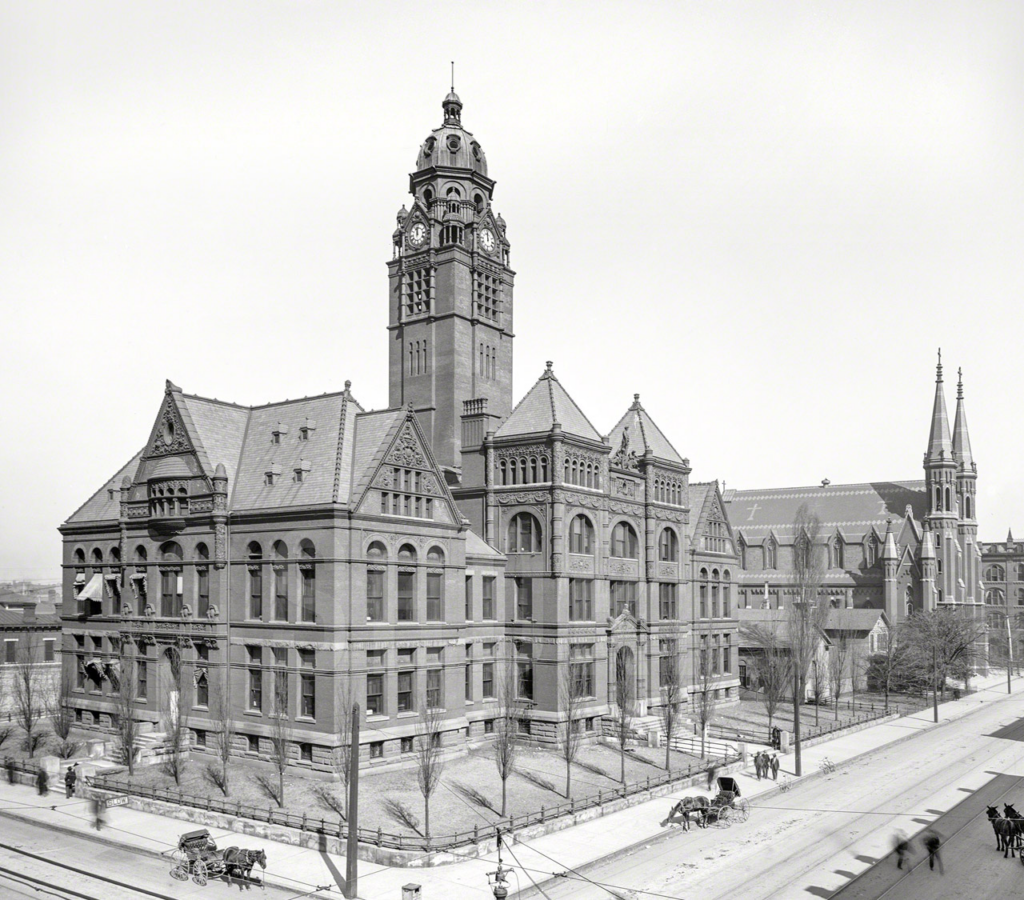 Historic brick building with a clock tower, featuring ornate architectural details, alongside a church with spires, captured in black and white.
