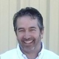 A smiling man with short, dark hair, wearing a white shirt, in front of a light-colored, textured background.